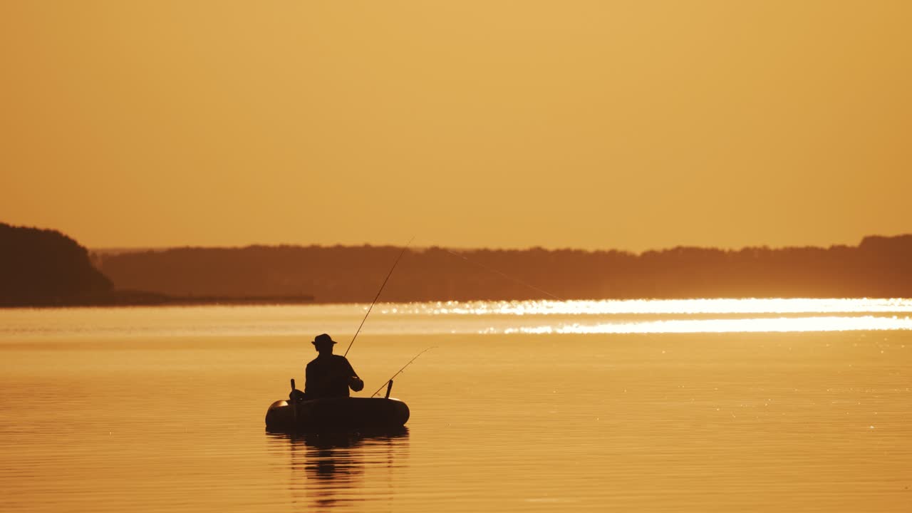 Silhouette of a man in hat fishing from the boat on the evening river background. Beautiful sunset and a fisherman catching fish from the inflatable boat in the open water.