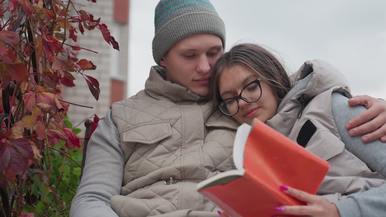 Beautiful cozy moment of young couple reading book together outdoors surrounded by autumn leaves sitting closely in warm jackets enjoying peaceful time with love and affection under cloudy sky