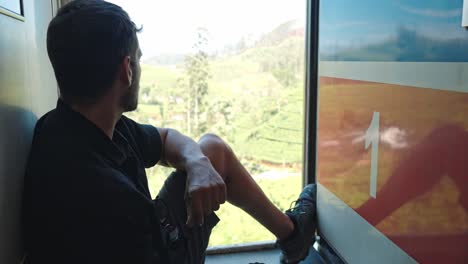 footage of a man sitting in the open doorway of a moving train, gazing out at the lush hills and tea plantations along the iconic ella–kandy railway in sri lanka.