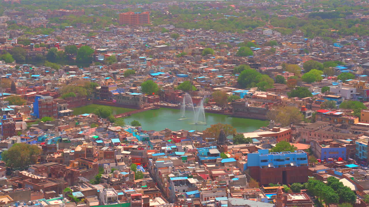 Una ripresa statica con teleobiettivo che ingrandisce il lago Gulab Sagar con le sue fontane decorative, circondato dalla densa architettura blu e colorata di Jodhpur, Rajasthan.