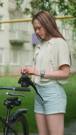 joven está de pie cerca de su bicicleta negra, se pone sus guantes en un entorno urbano pacífico, el fondo presenta un edificio residencial y un poste amarillo, rodeado de vegetación