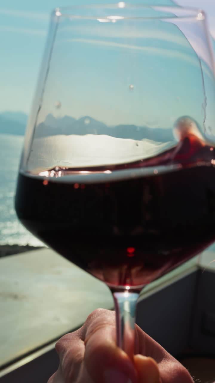 Close up of a woman swirling a glass of red wine on a table at a terrace with a sea view. Vertical