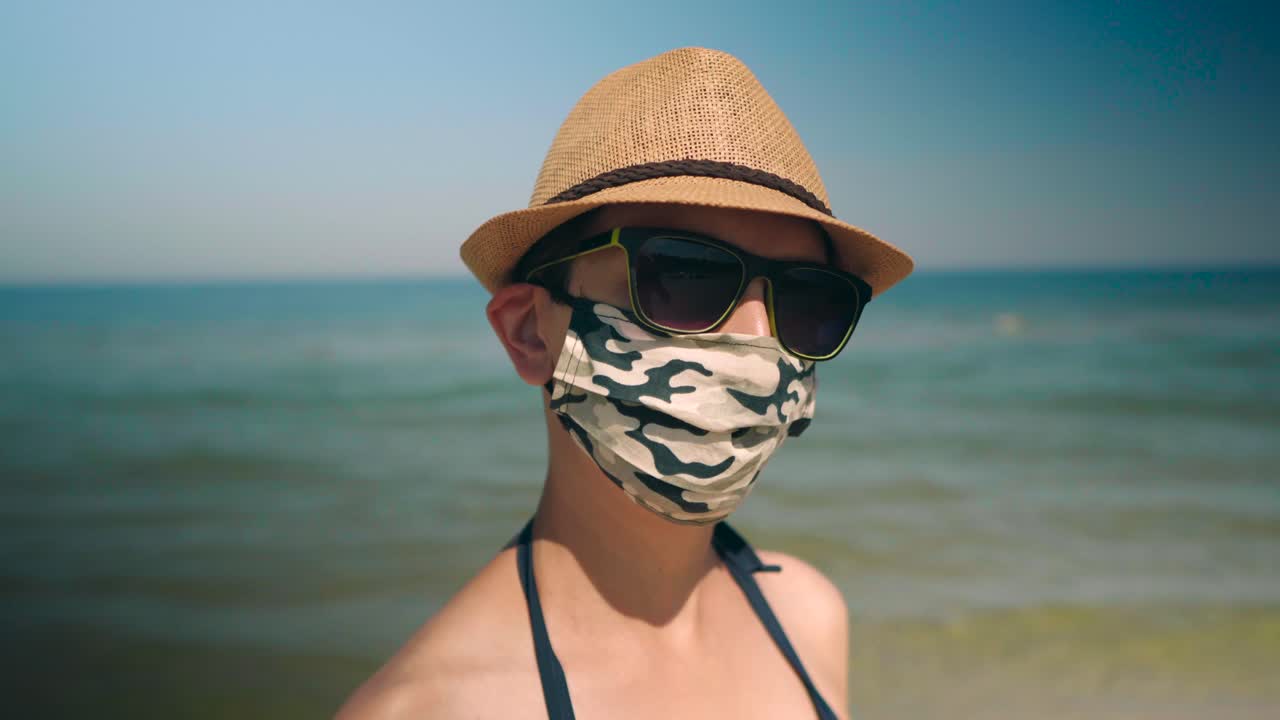 mujer usa máscara de corona con gafas de sol y sombrero en la playa al lado del océano