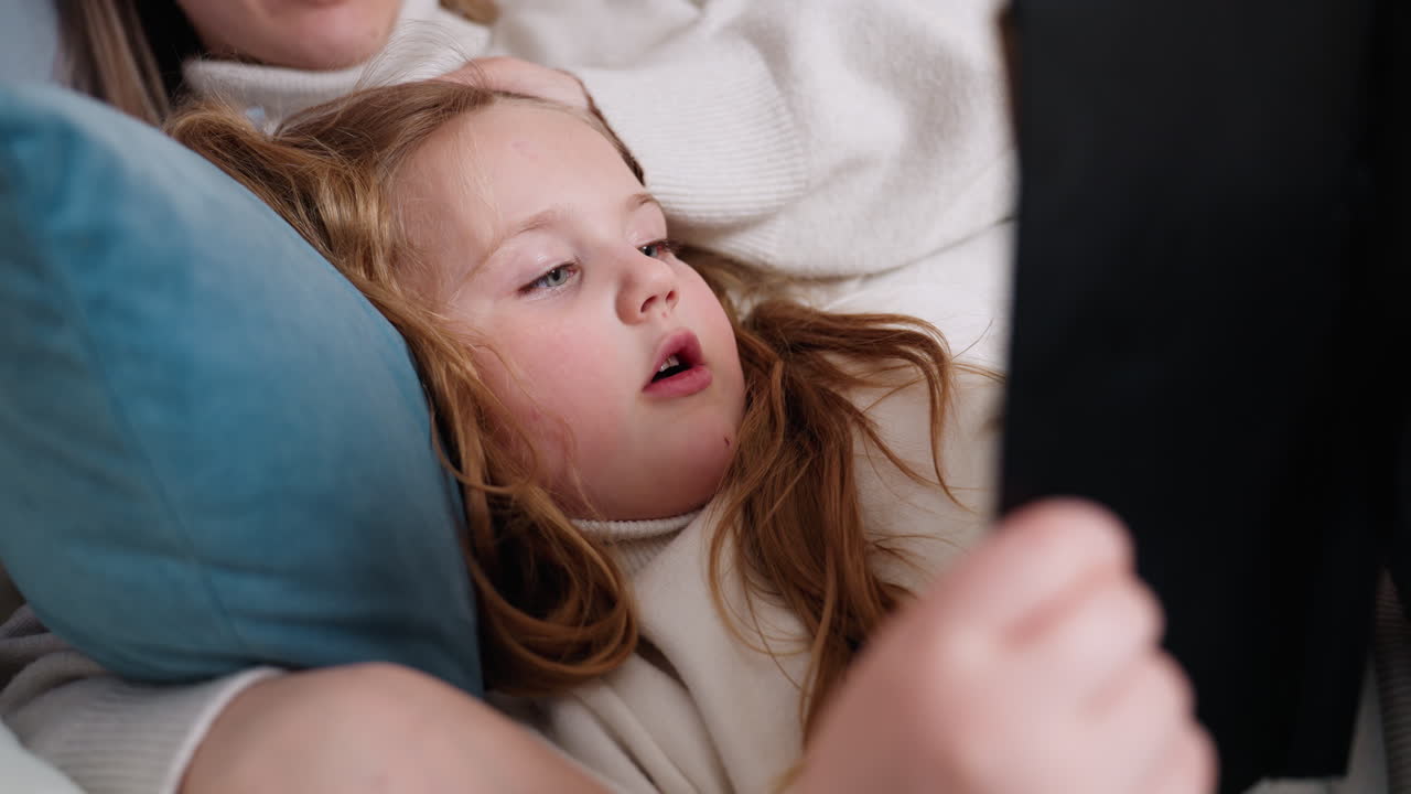 Young girl lying on bed reading book with mother beside, focused child turning pages while resting on pillows, cozy family bonding moment, peaceful learning and storytelling atmosphere in warm home