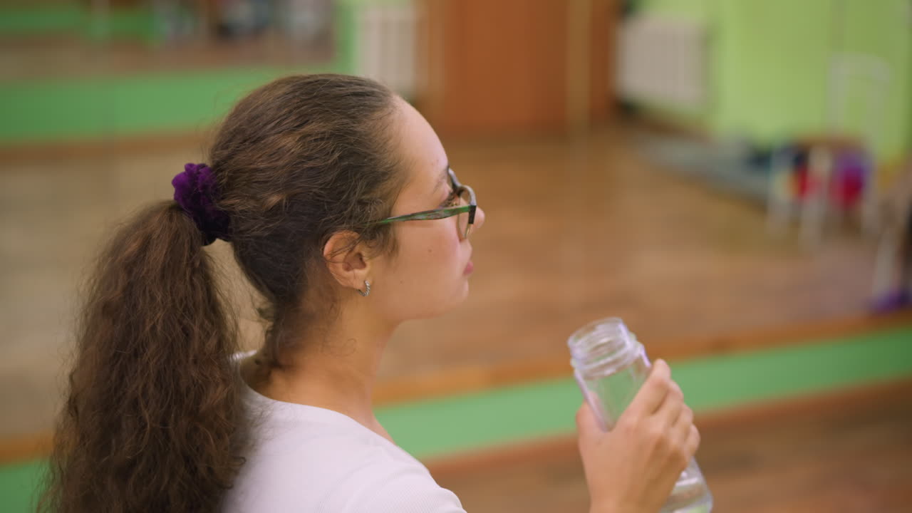Side view of woman in white top and glasses drinking water from clear bottle with relaxed posture, calm mood, and blurred bright studio background showing hydration and recovery after exercise