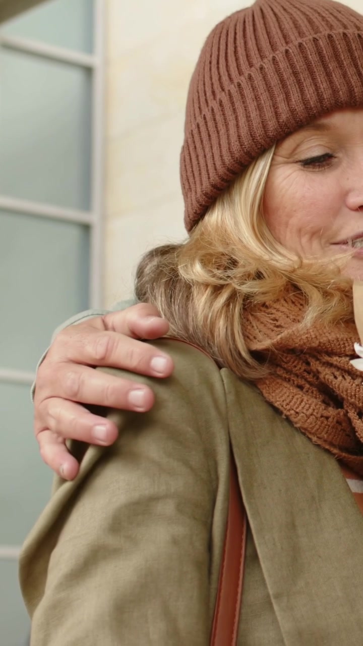 Video verticale: Un partner offre un bouquet a una donna con un cappello che lo riceve alla porta, abbracciandosi con affetto.