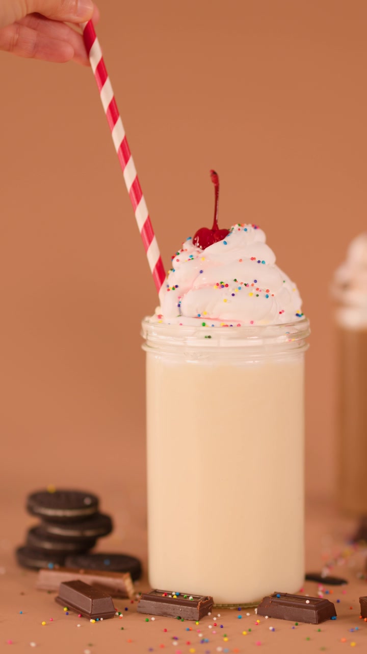 Close-up of hand inserting striped straw into creamy vanilla milkshake with whipped cream