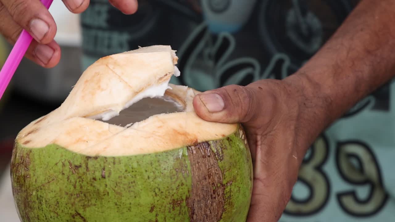 Close-up of a hand holding a green coconut with a straw inserted for drinking.