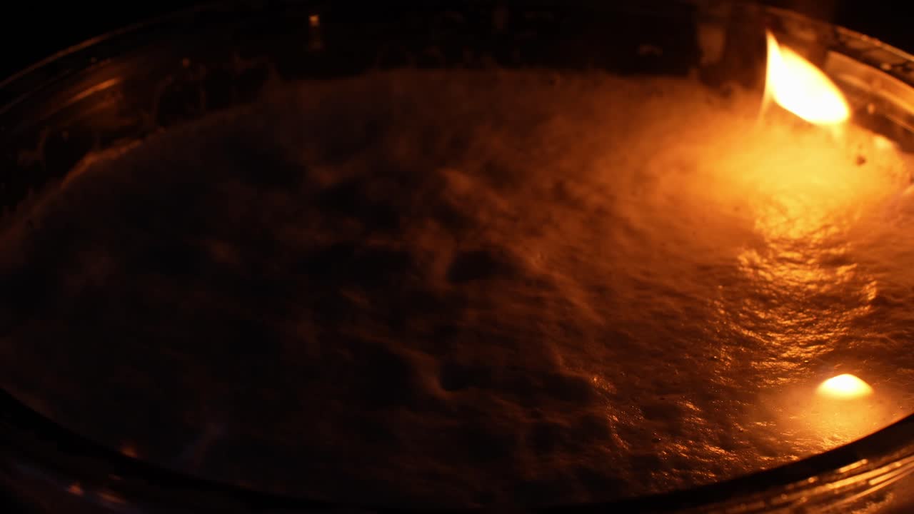 Flames dancing over a liquid surface within a glass container, creating a vibrant display of hot orange and yellow colors representing power, warmth, and intense energy, extreme close up, macro
