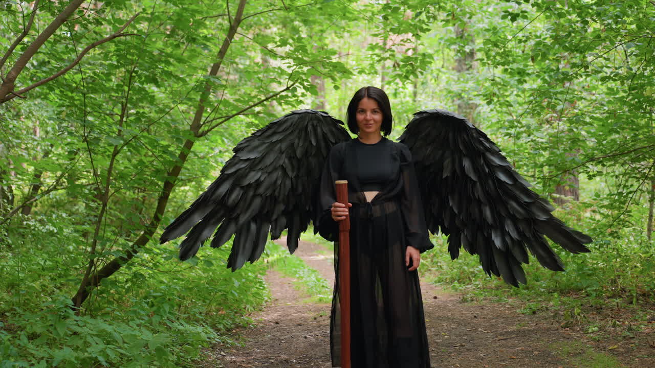 Young woman dressed in black feather costume giving soft air kiss with gentle smile in lush green forest path, holding wooden staff, surrounded by sunlight and foliage