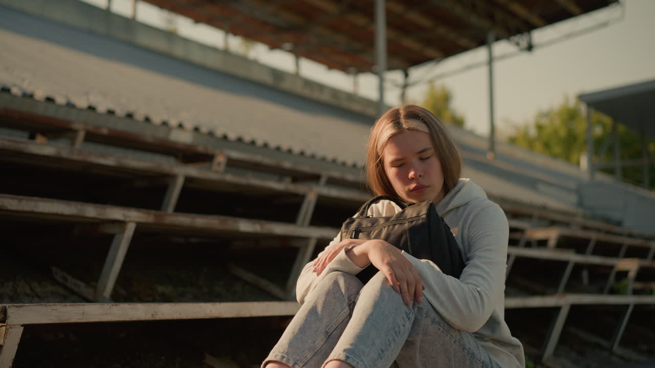 Close-up of a young woman gazing thoughtfully while seated alone on empty stadium bleachers, hugging her bag tightly with a reflective expression, sunlight softly illuminates her face