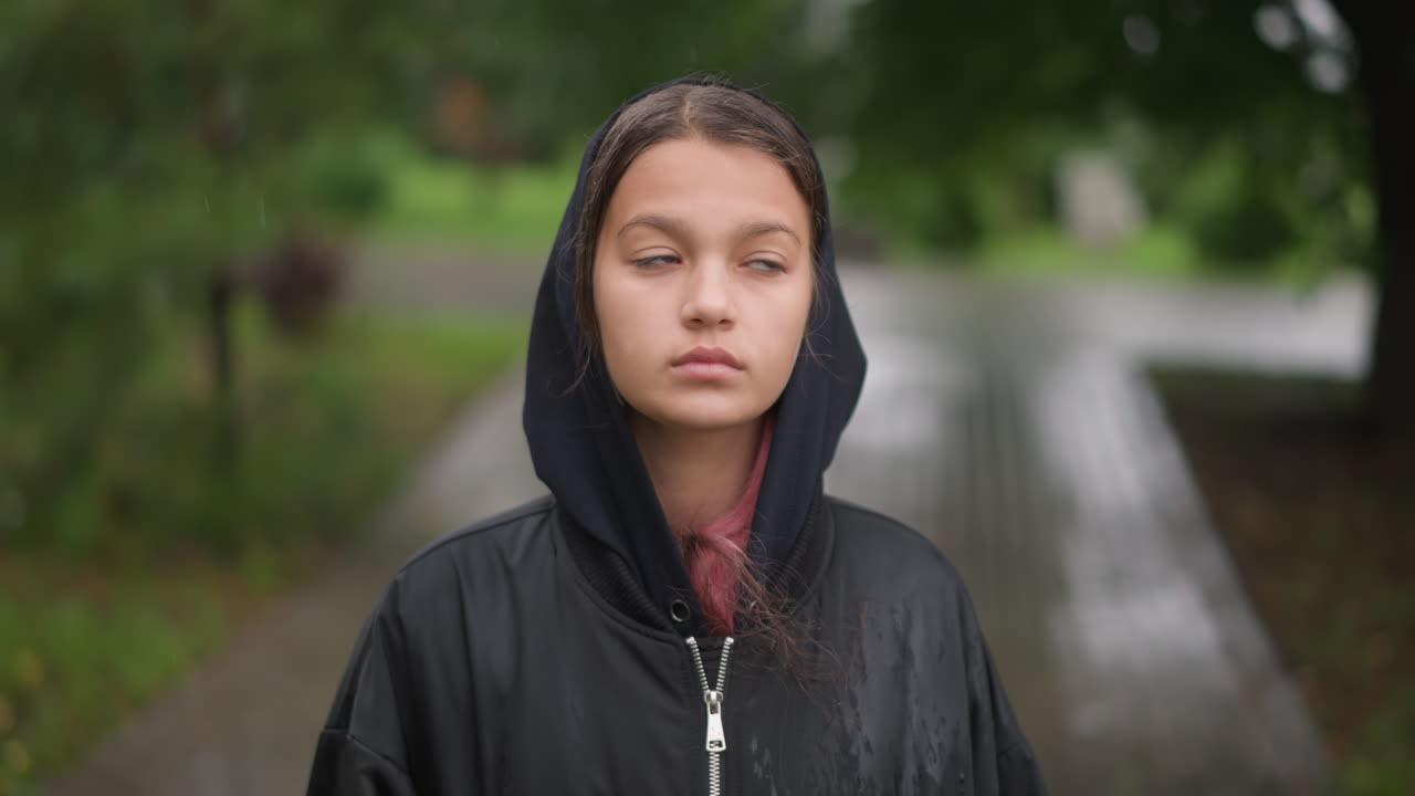 Ung kvinne ser opp gjennom lett yr på regnvåt brygge, dråper klamrer seg til hetten mens trekronen rammer inn en myk himmel, håpefull, ettertenksom stemning med filmatisk lys og en øm følelse.