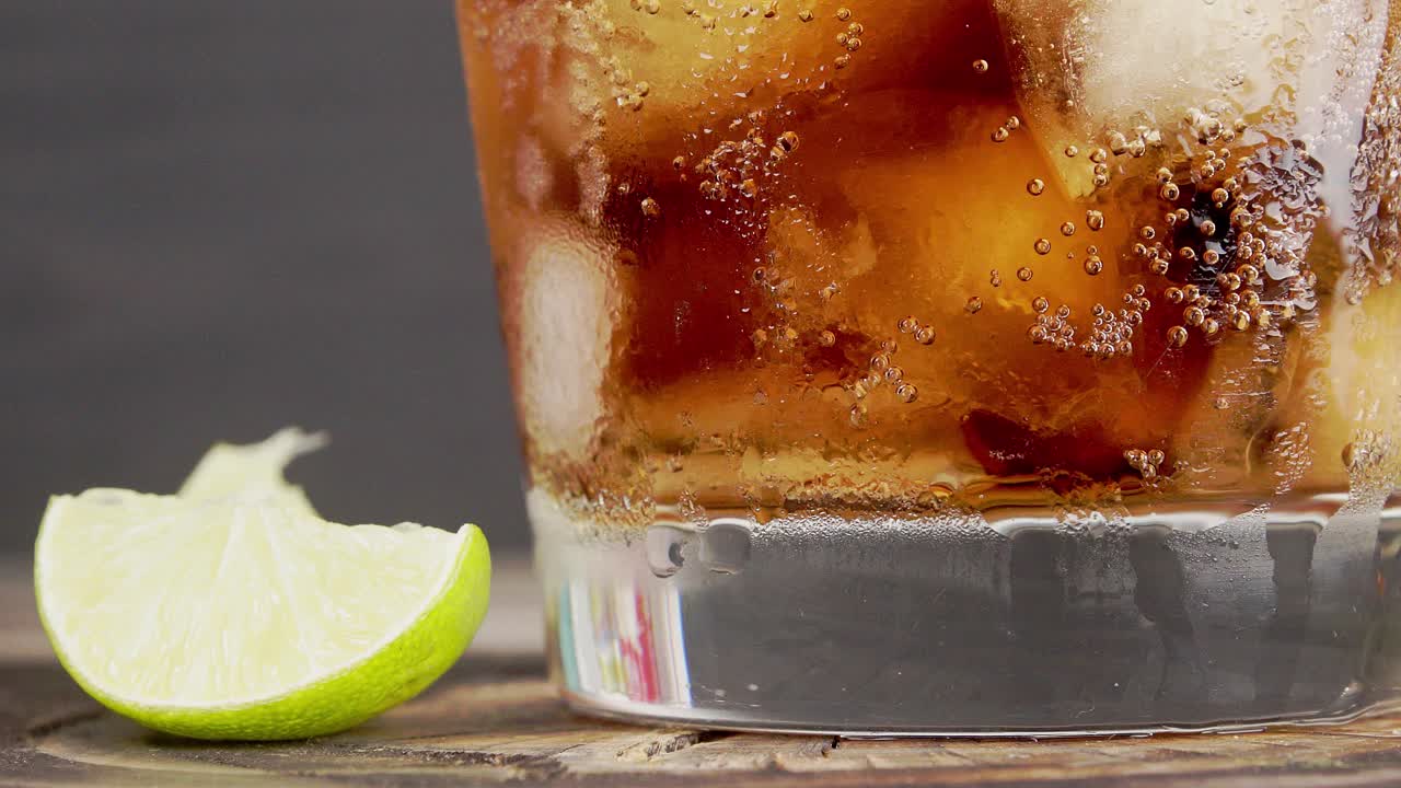 Glass of cold soda with ice cubes and fizzy bubbles, lemon slice aside on a wooden table - static close up  shot