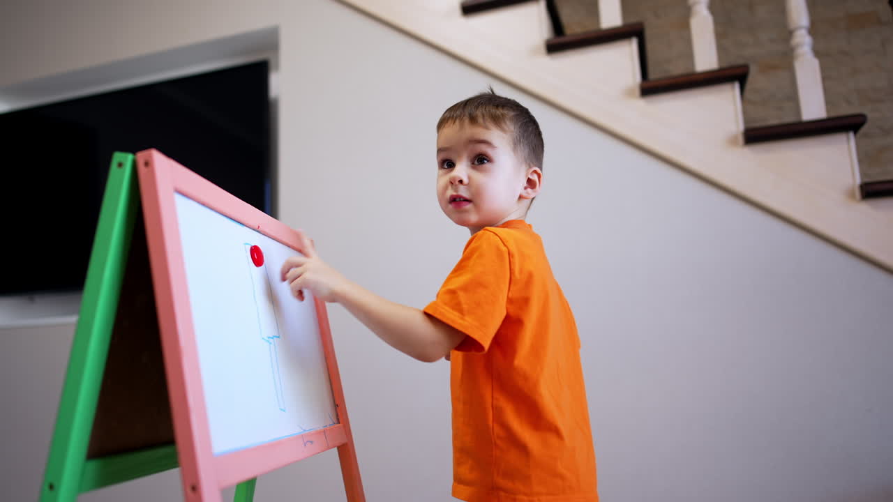 Lovely baby boy in orange t-shirt stands at the whiteboard. Cute kid points at the red magnet on the board.