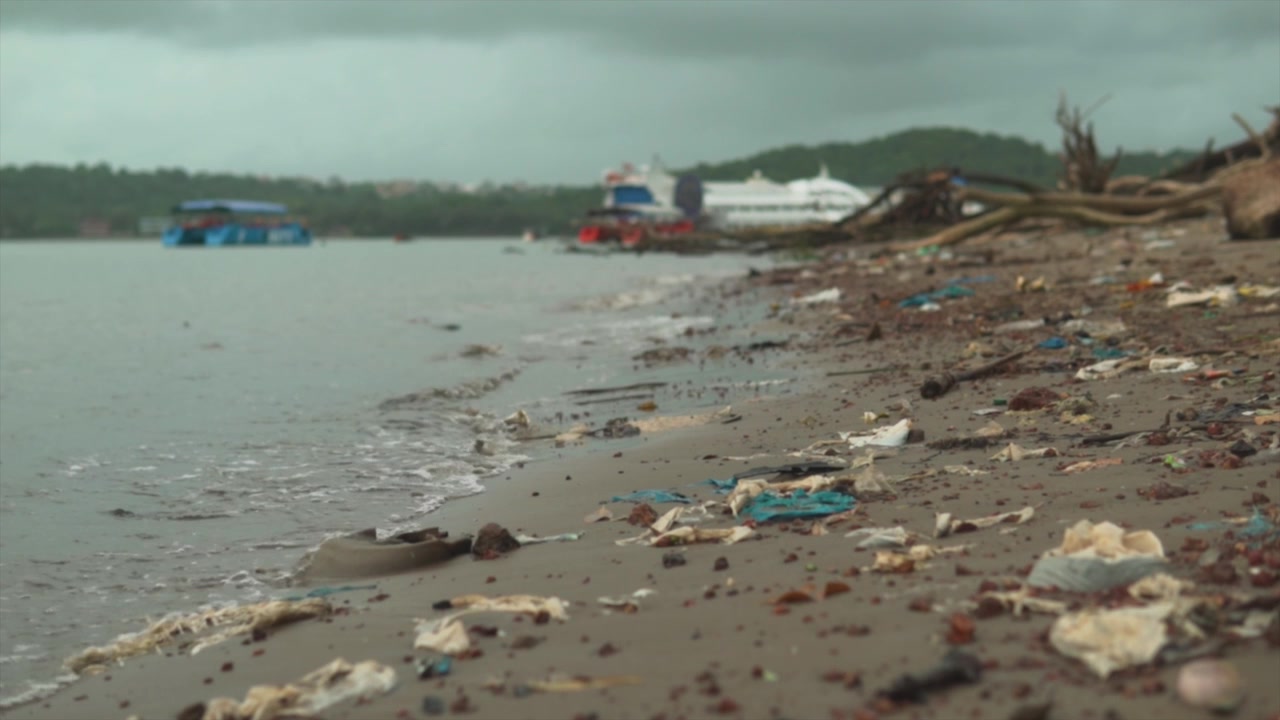In a slow-motion shot with a shallow depth of field, the Indian shoreline is sadly marred by litter, while boats can be seen in the distance