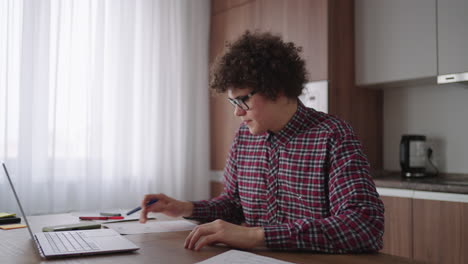 curly haired male student attractive young boy in glasses is studying at home using laptop typing writing in notebook. college student using laptop computer watching distance online learning seminar