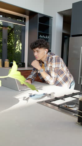 vertical video: young man using laptop in modern kitchen, listening with wireless earbuds
