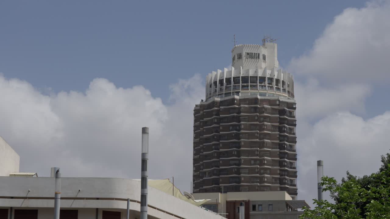 Tall building in Dizengoff, Tel Aviv, Israel with lower building in foreground. Blue skies with clouds. Bird flying. Wide shot. Slight pan right
