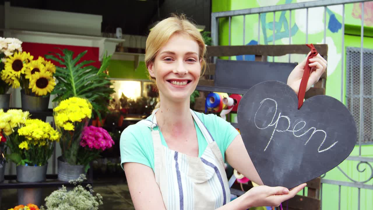 Lächelnde Floristin hält ein Geöffnet-Schild auf einer Schiefertafel im Blumengeschäft.
