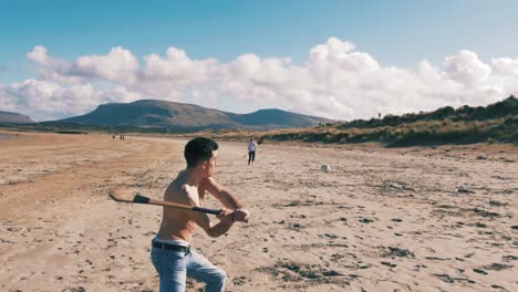 joven lanzando y golpeando una pelota de hurley con un palo de hurley en la playa con un hermoso paisaje montañoso en el fondo