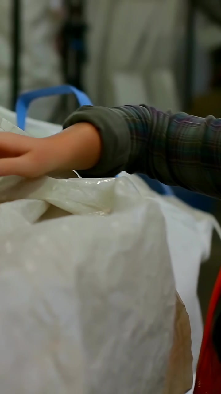 Video verticale: Operaio con gilet fluorescente che ispeziona, si allunga e solleva della plastica in un negozio, esponendo della segatura.