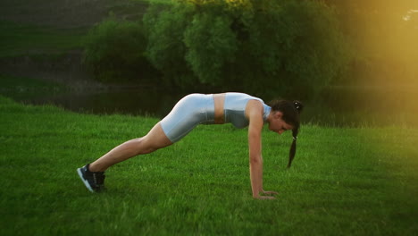 a woman performs a plank exercise standing on the grass at sunset in a park. slowly goes on the hands of on the grass. lift your legs in the plank exercise.