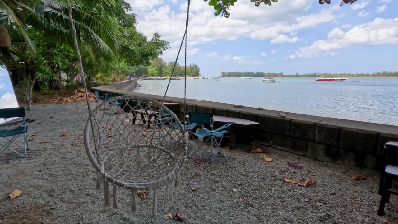 Empty rope swing sways on shaded pebbled beach, calm water and blue sky beyond