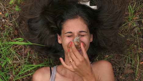 a pretty brunette woman laying in the grass in a forest while placing a snail on her nose and acting grossed out and uncertain