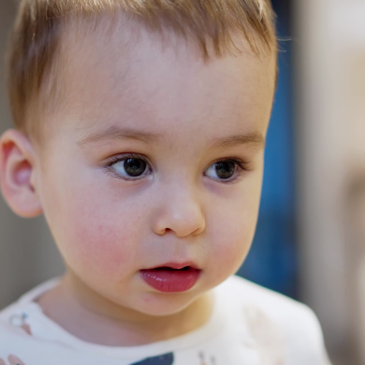 Beautiful Caucasian baby portrait. Adorable toddler boy sits calm smiling to the camera and approaching it. Close up. Blurred backdrop