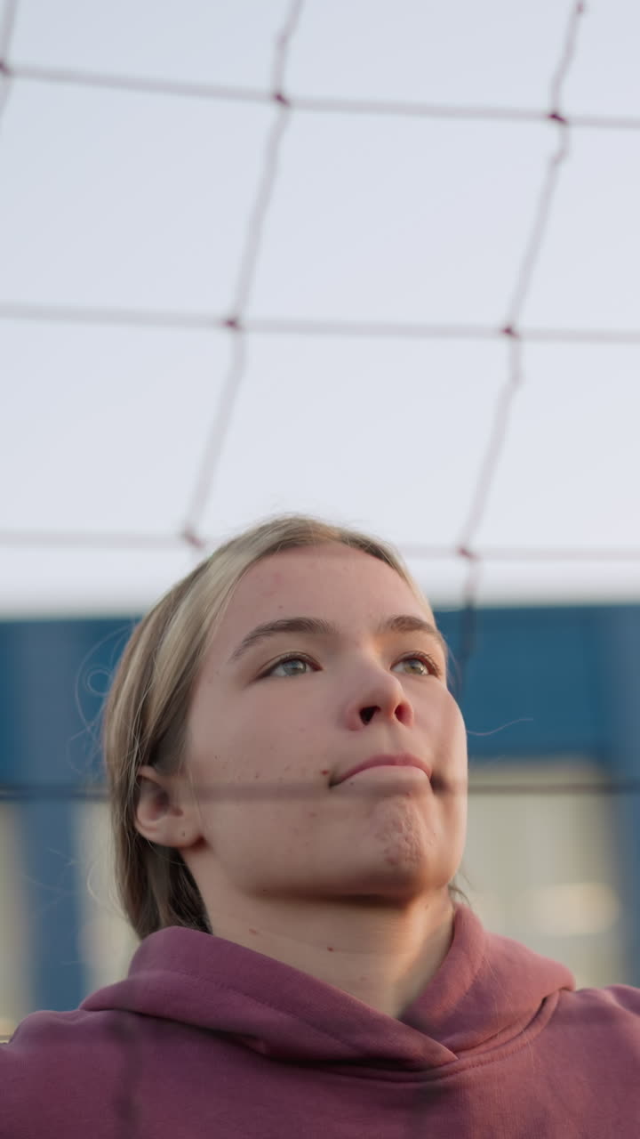 primer plano de una joven atleta que salta y se prepara para golpear el voleibol, con enfoque en su expresión y determinación, el fondo presenta una red de voleibol y edificios