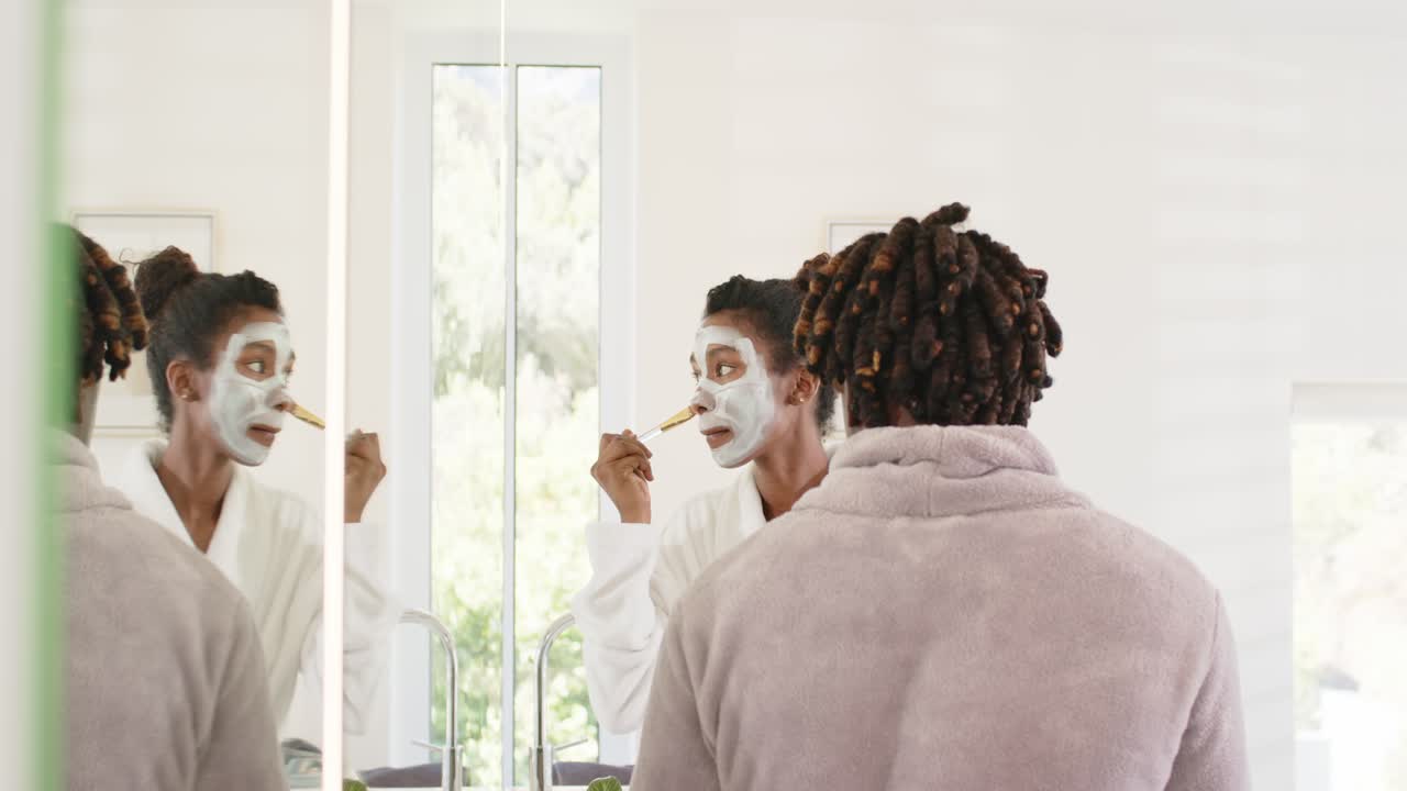 Pareja afroamericana en el lavabo, mujer sosteniendo un cepillo aplicando una mascarilla blanca revisándose en el espejo, hombre de espaldas