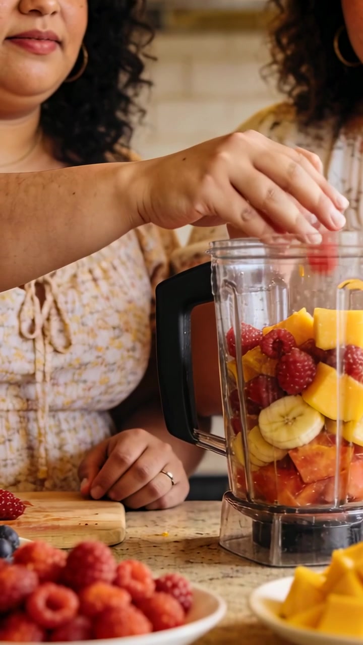 Video verticale: Due donne con maglie a fantasia che aggiungono frutta nel frullatore sul bancone per una bevanda.
