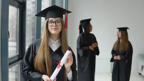 the graduate holds a diploma in one hand and a hat in the other. master's degree in the university on the background of two female students of african american and caucasian races