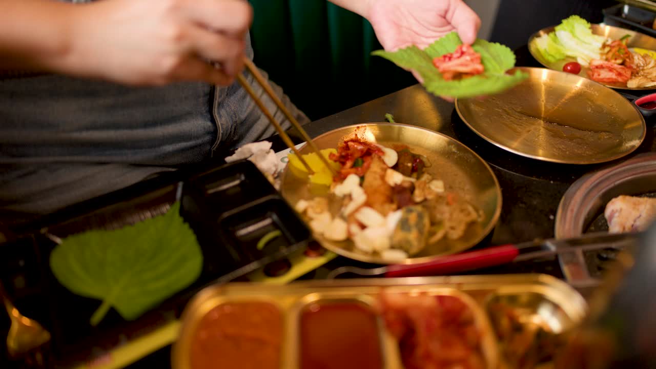 Person uses chopsticks to add pork and kimchi to perilla leaf at Korean barbecue table
