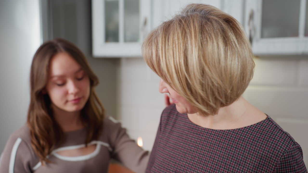 Bedstemor retter håret, mens hun varmt og kærligt ser på barnet. Den hyggelige køkkenkulisse med slørede skabe i baggrunden giver en hjemlig følelse
