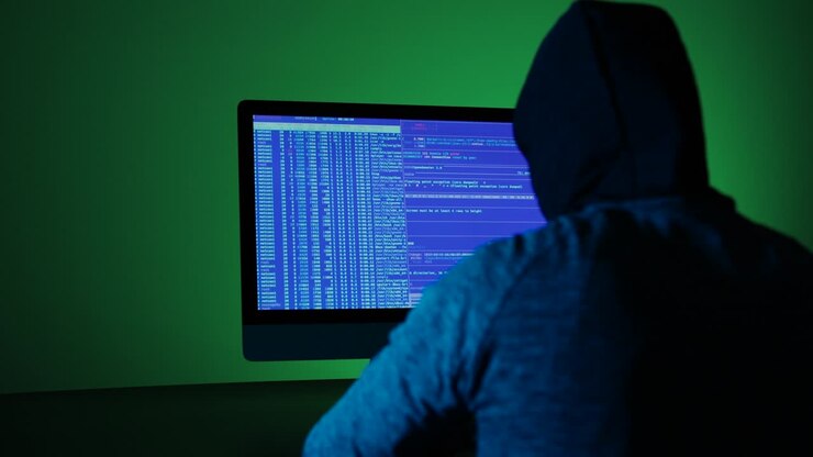 Vista posteriore di un programmatore hacker con cappuccio, che scrive codice verde su uno schermo di computer. Uomo irriconoscibile che hackera in una stanza buia