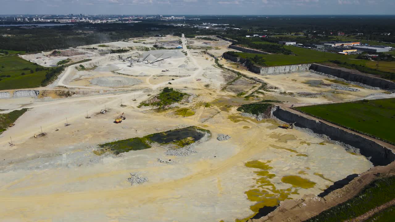 Aerial drone flyover shot of a large limestone quarry mine with excavators and conveyor belts visible in a dusty dug pit in green countryside nature during a summer sunny day. Waste water puddles also