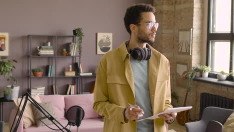 hombre con auriculares de pie sosteniendo papeles en una habitación con micrófono y mirando a la cámara 1