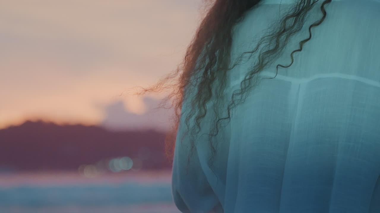 Woman watching sunset. Distant island scene highlighted by warm light. Curly haired woman gazes at sunset with windblown hair. Woman with curly hair observes sunset as wind blows her hair around