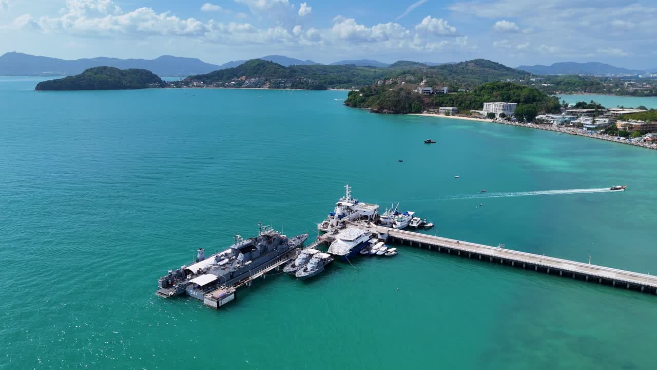 Drone footage captures military boats docked at a pier in Phuket, Thailand, surrounded by turquoise waters and lush landscapes