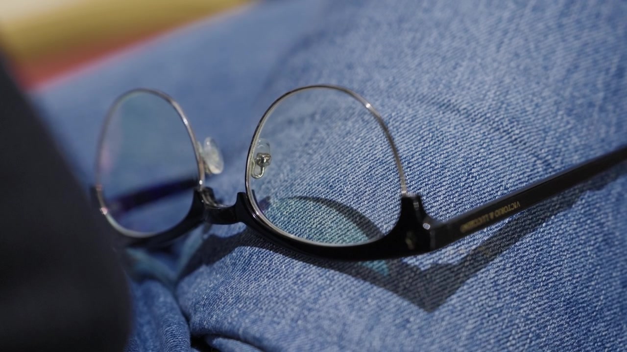 Kacamata bingkai kawat bulat dengan lensa anti cahaya biru bertumpu pada kain denim biru terlipat dalam cahaya lembut