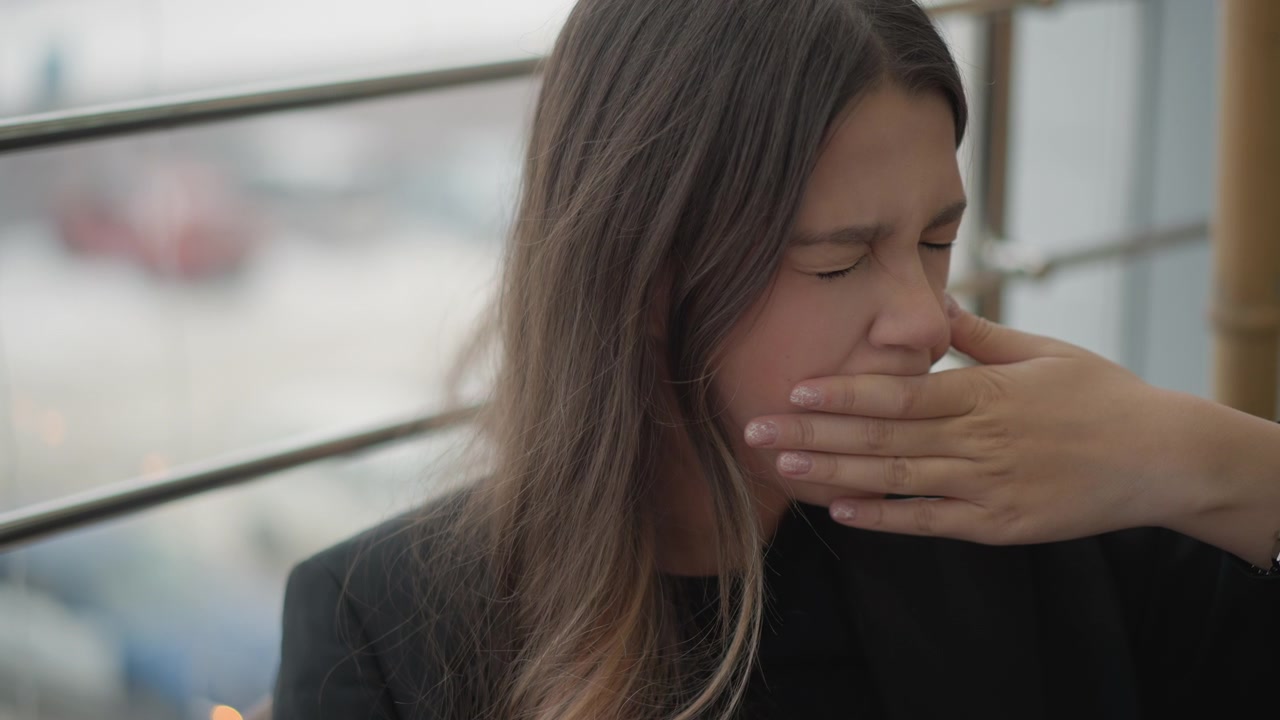 Vista ravvicinata di una giovane donna seduta accanto a una finestra con ringhiera urbana, che gira la testa di lato e si tocca la bocca mentre guarda in basso, mostrando quieta contemplazione, sottile emozione, luce diurna soffusa e spontaneità.