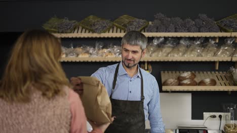 Friendly-smiling-male-cashier-in-apron-giving-paper-bag-with-bread-to-the-smiling-woman-and-she-leaves-holding-paper-cup-with