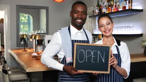 smiling waiter and waitress holding slate with open sign in restaurant