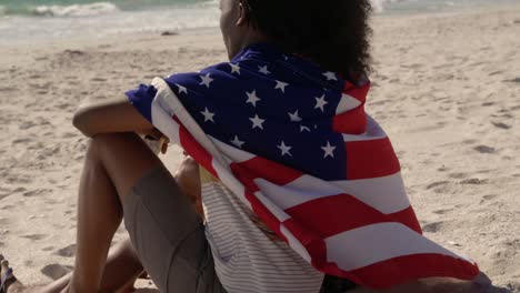 Seitenansicht-Eines-In-Amerikanische-Flagge-Gehüllten-Afroamerikanischen-Paares,-Das-Zusammen-Am-Strand-Sitzt-4k