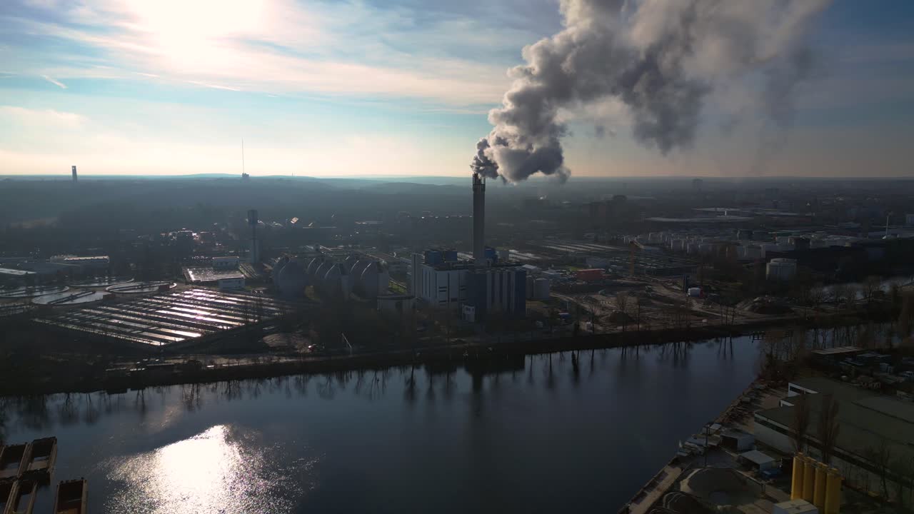 large industrial power plant emitting large amounts of smoke over the city Berlin next to a river on a sunny day. Fabulous aerial view flight static tripod hovering drone