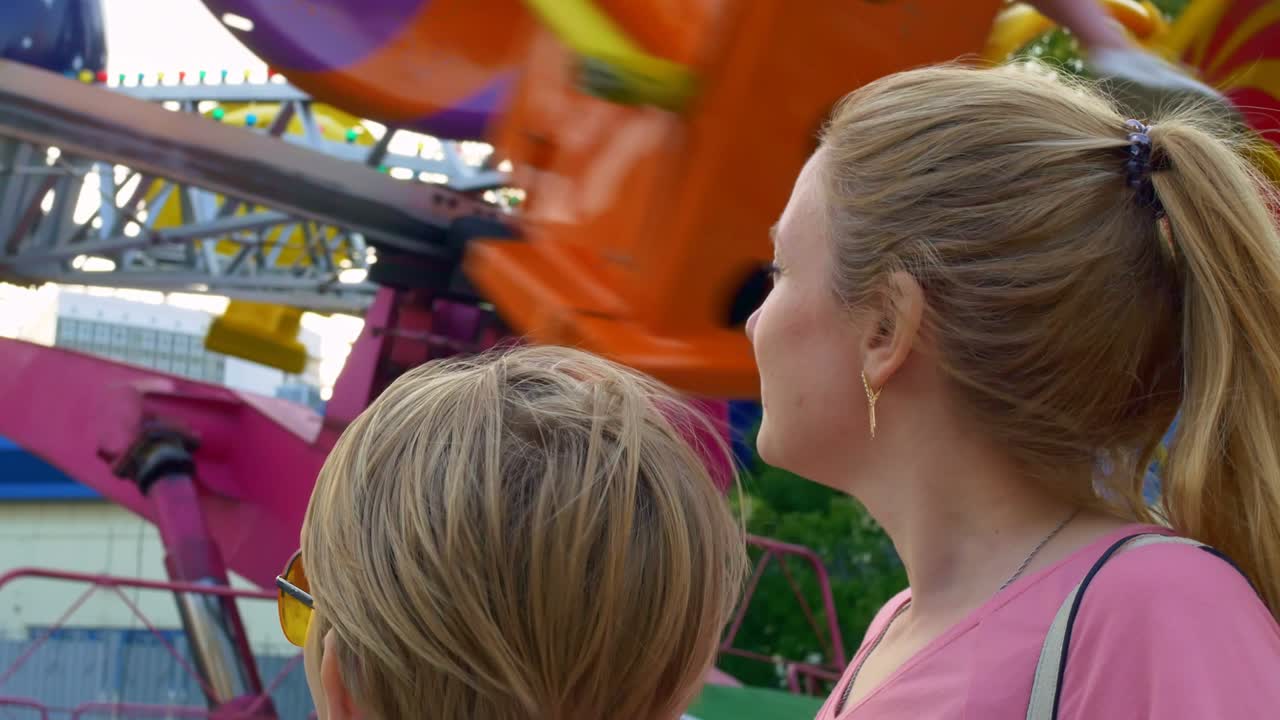 primer plano de una madre y un hijo en un parque de atracciones, de pie junto a un carrusel que gira muy rápido, cabello rubio revoloteando en el viento. infancia alegre en la feria del domingo