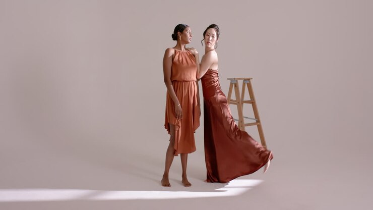 Diverse modelle con la mano appoggiata sulla spalla e in posa mostrando abiti color ruggine in studio vicino a una scala.