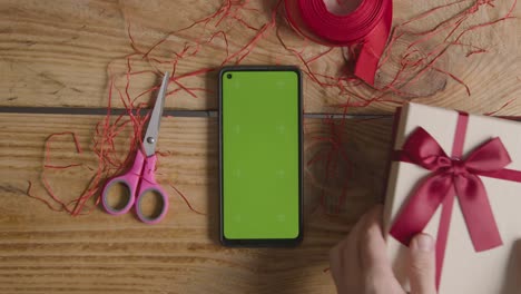 fotografía cenital de un hombre que envuelve un regalo de san valentín romántico presente en una caja junto a un teléfono móvil con pantalla verde