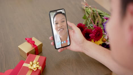 Mujer-Caucásica-Sosteniendo-Un-Teléfono-Inteligente-Con-Una-Mujer-Birracial-En-La-Pantalla,-Regalos-En-El-Escritorio